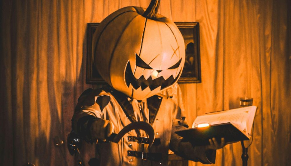 A creepy character in a pumpkin mask holds an illuminated book in one hand, and a sickle in the other.