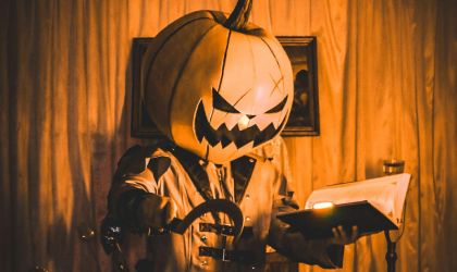 A creepy character in a pumpkin mask holds an illuminated book in one hand, and a sickle in the other.