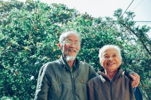 an elderly couple posing and smiling in front of a hedge.