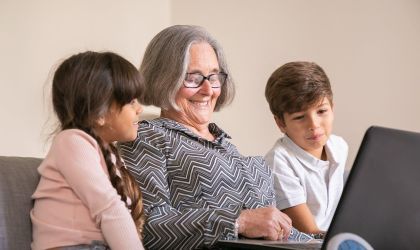 A woman sits on the couch using a laptop and two children look on